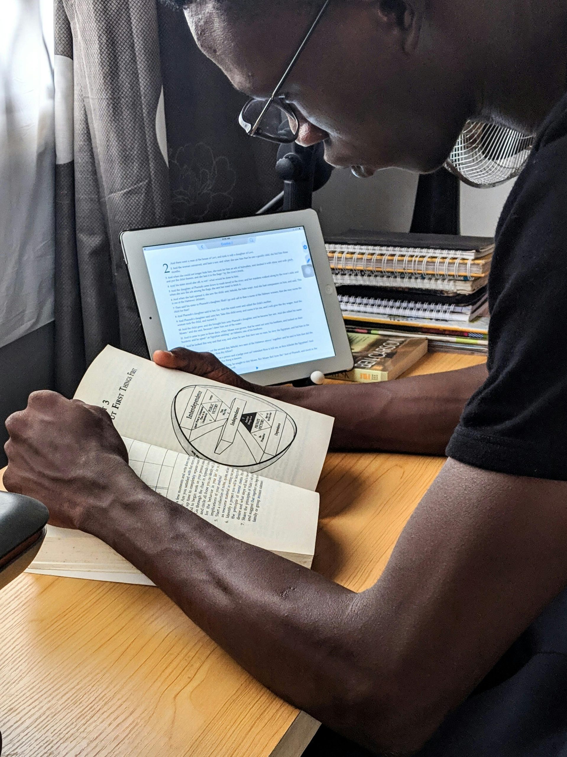 A man studying with a book and tablet at a desk indoors, showcasing multitasking and modern study habits.